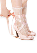 A person wearing pale pink satin high-heeled shoes with lace socks and long ribbon ties, shown from the hands down against a white background.