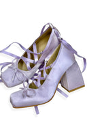 A pair of lavender satin high-heeled shoes with matching ribbon laces and bows, displayed on a clean white background.