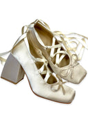 A pair of women's block-heel mules in a light champagne satin fabric, featuring square toes and delicate ribbon laces that wrap around the instep and tie at the toes, set against a plain white background.