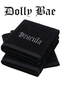 Three folded black towels stacked on a white background, with the top towel embroidered with the word 'Marcula' in a gothic font. The text 'Dolly Bae' appears above in the same gothic typeface.