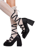 Black high-heeled platform shoes with large bows and wraparound lace-up straps, shown on a model wearing cream ribbed knee-high socks against a white background.