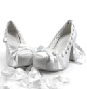 A pair of white satin high-heeled shoes with ribbon bows and lace-up fronts, displayed on a white background with the text 'Dolly Bae' above them.