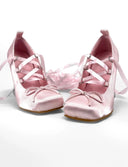 A pair of light pink satin Mary Jane shoes with ribbon laces and bow details, displayed on a white background with the text 'Dolly Bae' above them.