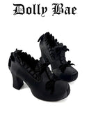 A pair of black gothic-style platform heels with ruffled trim and bows, shown on a plain white background with the brand name 'Dolly Bae' printed above in a black gothic font.