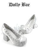 A pair of white satin high-heeled shoes with lace-up front details and ribbon bows, displayed on a white background with the text 'Dolly Bae' above them.