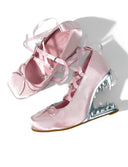 A pair of light pink satin wedge shoes with ribbon laces and a metallic spiked heel, shown on a white background with the text 'Dolly Bae' above them.