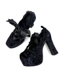 A pair of black lace-up high-heeled shoes with a textured velvet-like finish and large black ribbon bows, shown on a plain white background with the text 'Dolly Bae' above them.