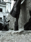 A gothic-style fashion scene featuring a model wearing black fishnet stockings and high-heeled boots, standing on a stone ledge in front of a castle. A black birdcage with a bird sits to the left, and a black rabbit figure is visible on the right.