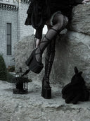 A person in a gothic outfit with black fishnet stockings, platform heels, and a dark dress sits on a stone wall beside a small black rabbit figurine and a birdcage in a stone courtyard.