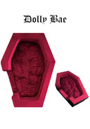A gothic-style coffin-shaped product shown from two angles on a plain white background, with the text 'Dolly Bae' above. The item appears to be a plush or padded coffin-shaped cushion/seat in a vivid pinkish-red tone with darker embroidered detailing inside.